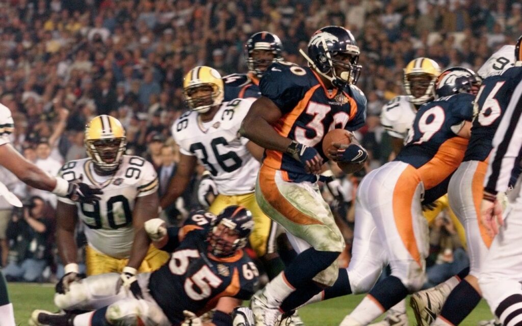 Terrell Davis runs a touchdown in against the Green Bay Packers in Super Bowl XXXII. He has on a navy blue and orange jersey with white pants. The Packers wore their white jerseys and yellow pants with green stripes.