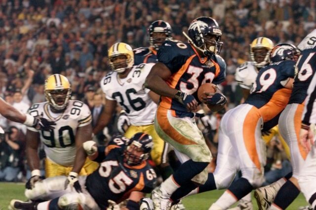 Terrell Davis runs a touchdown in against the Green Bay Packers in Super Bowl XXXII. He has on a navy blue and orange jersey with white pants. The Packers wore their white jerseys and yellow pants with green stripes.